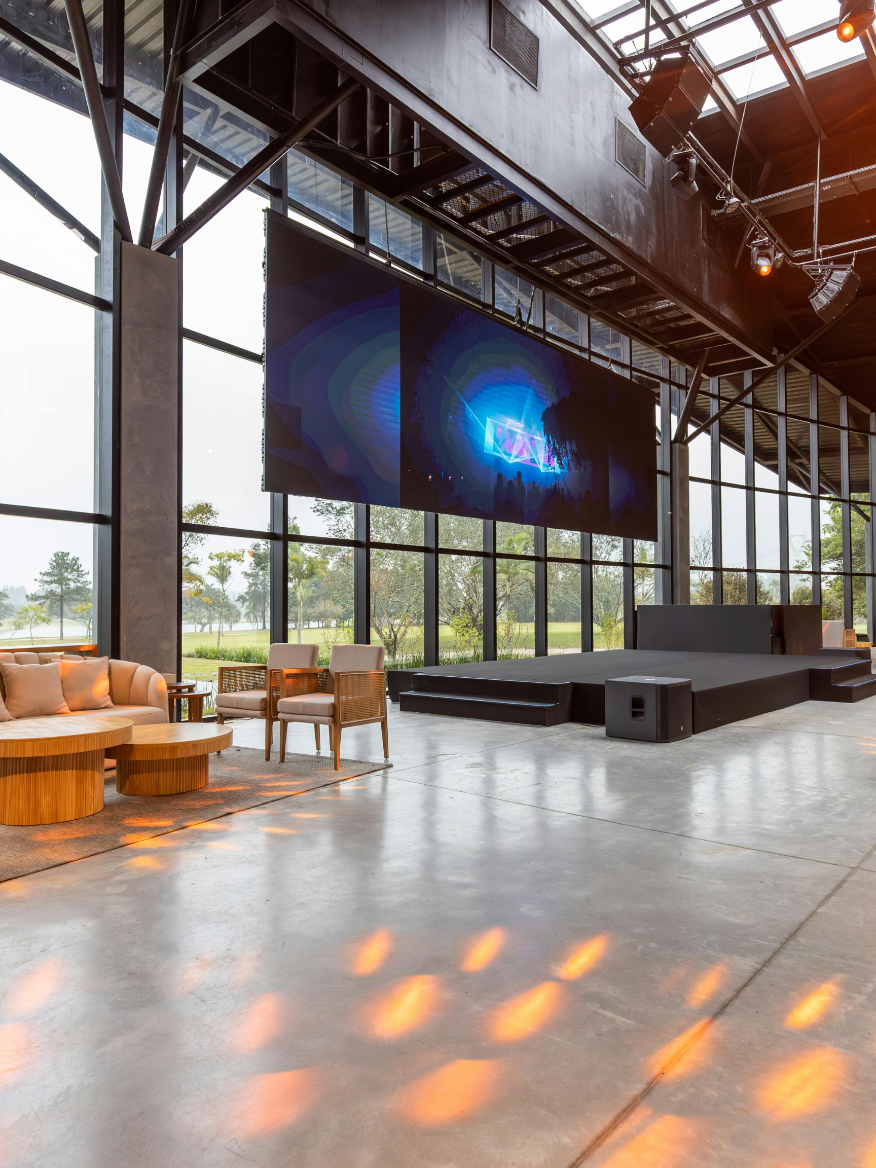Large indoor space with a stage, oversized window walls showing a green outdoor area, and seating including a cream sofa and wooden chairs. A large digital screen is above the stage, and a speaker is positioned at the front of the stage. Soft orange light reflections are on the polished concrete floor.