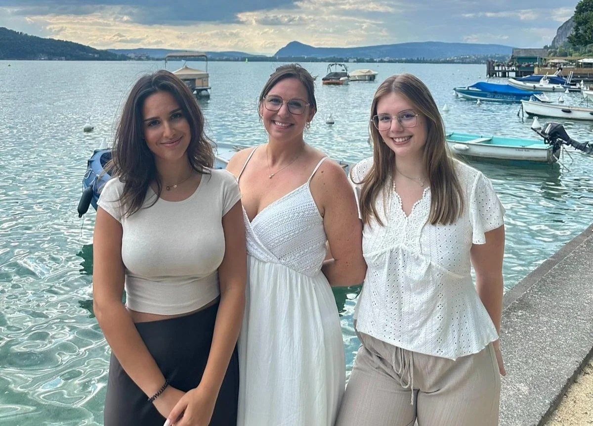 Trois femmes souriantes se tiennent près d'un lac avec des bateaux à moteur et une montagne en arrière-plan.