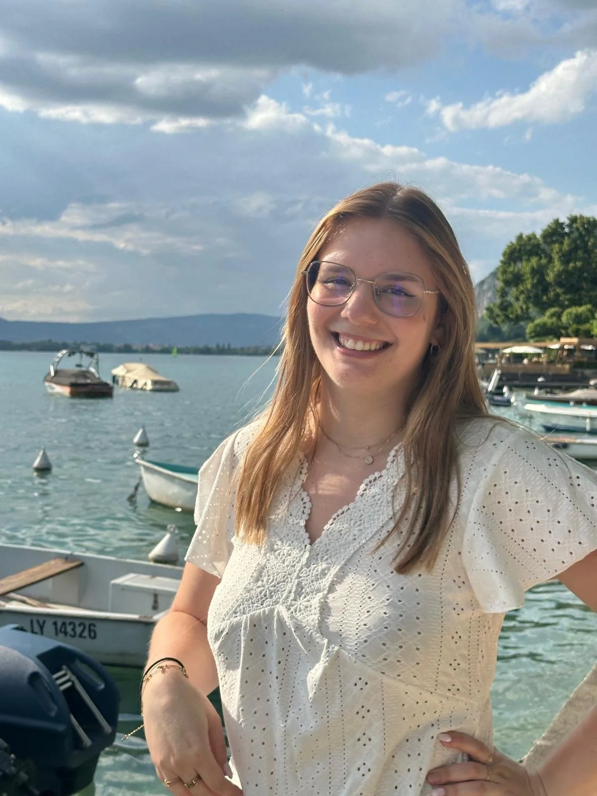 Jeune femme souriante avec lunettes, portant une blouse blanche, près d'un lac avec des bateaux et un ciel nuageux.