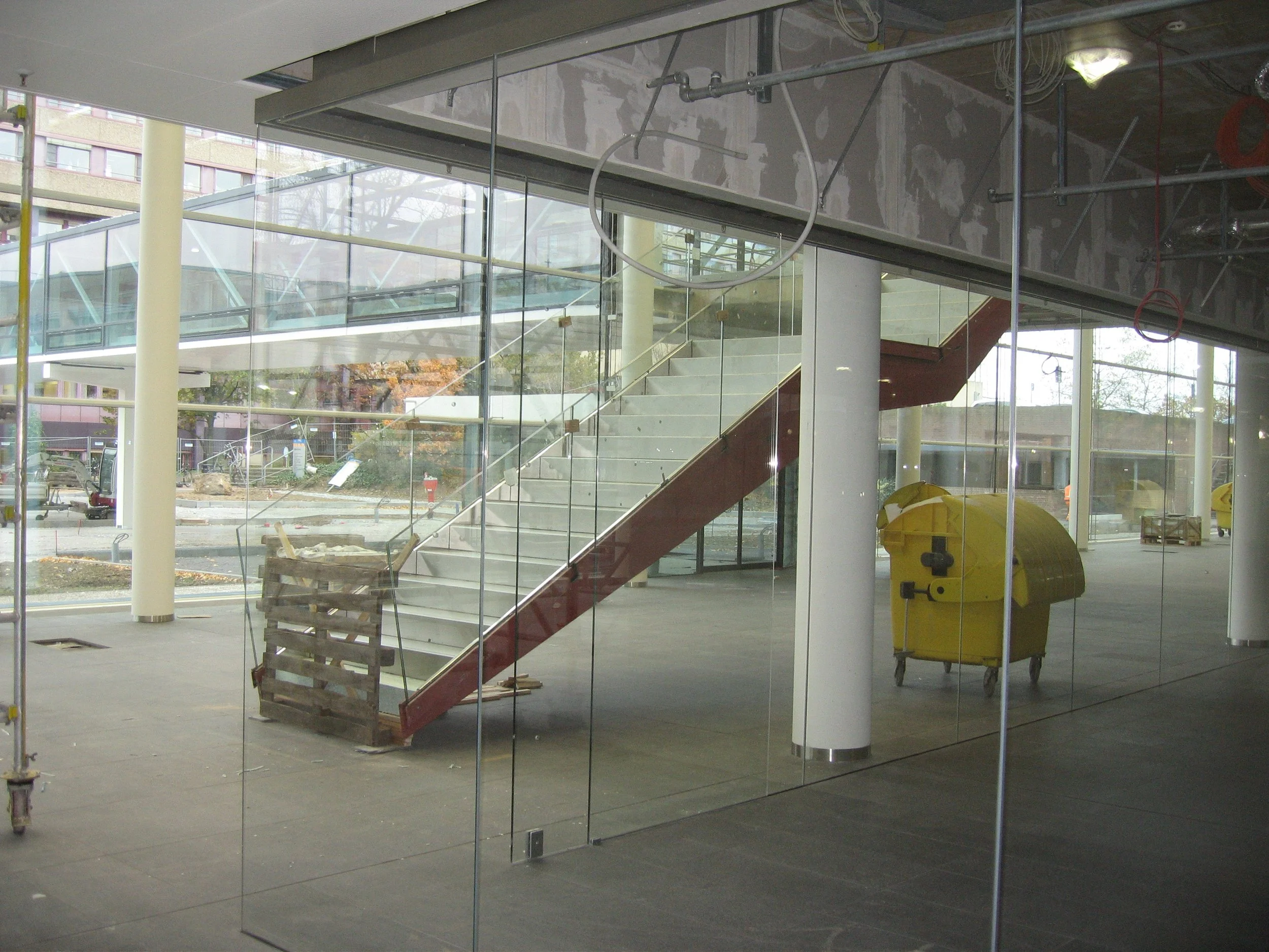 Innenansicht eines Gebäudes im Bau mit Glastrennung, Treppe, gelbem Mülleimer auf Rollen und Baugerüsten.