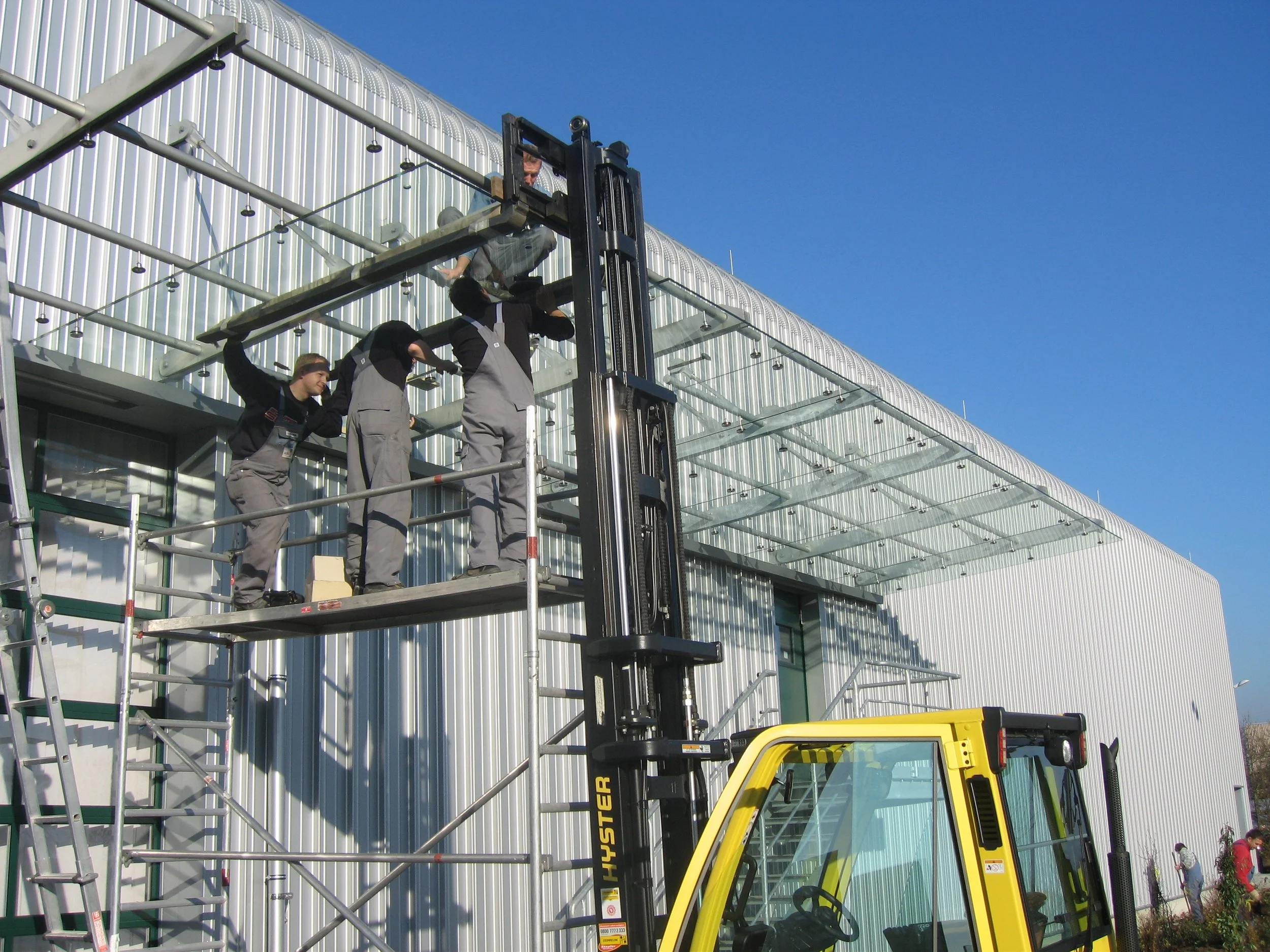 Drei Arbeiter auf einer Gerüsttreppe, die an einem modernen Gebäude mit einer Glasüberdachung arbeiten, während ein Gabelstapler daneben steht.