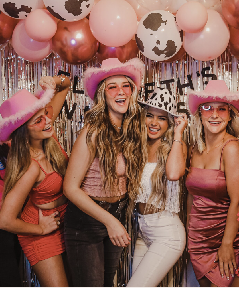 Four women celebrating in front of a festive backdrop with balloons and metallic streamers, wearing pink and cow print hats with sunglasses, smiling and laughing.