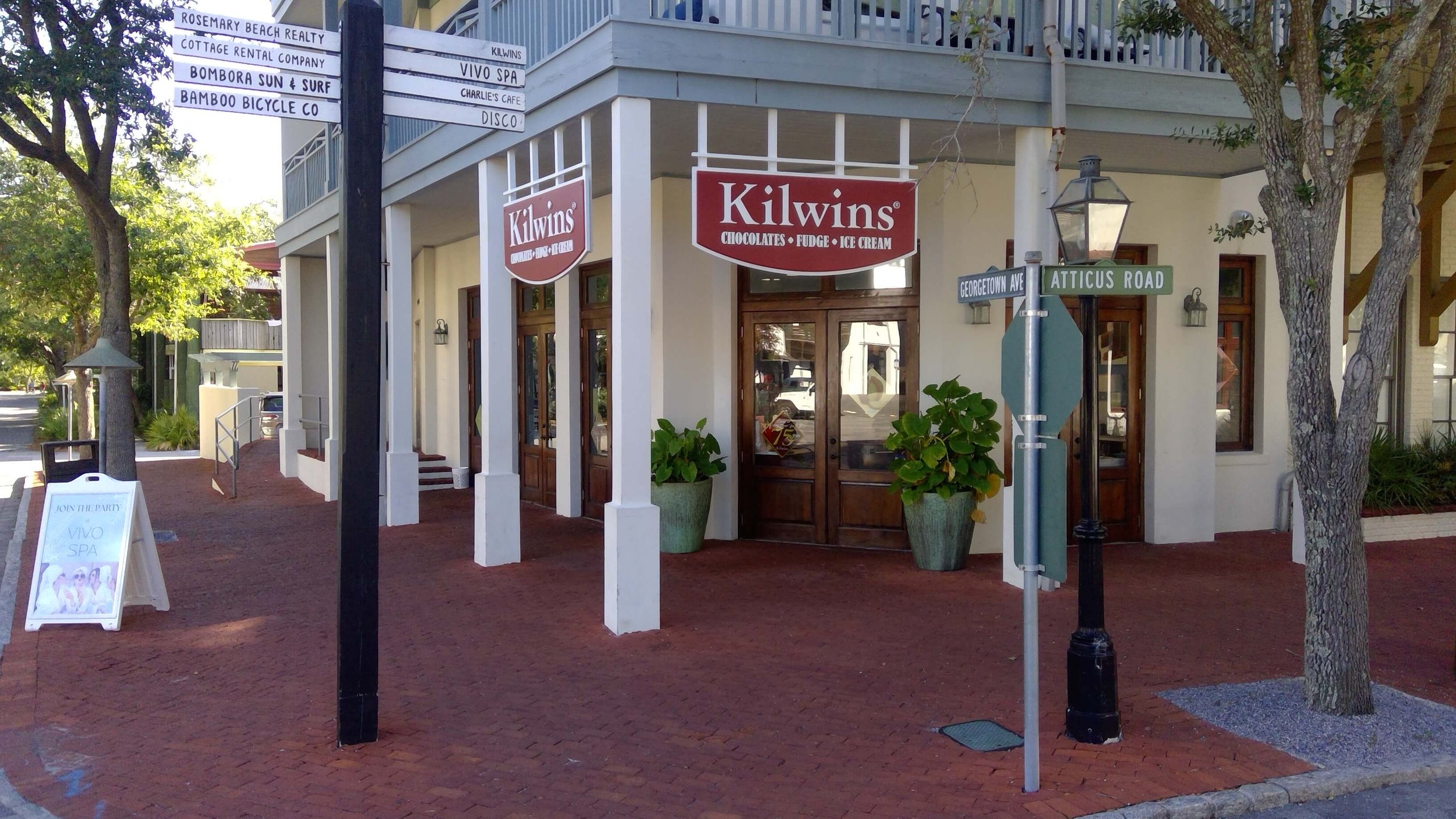 Corner of a building with signs for Kilwins, a street sign at Georgetown Avenue and Atticus Road, and a sidewalk with a white sandwich board for Vivo Spa. Trees and a street are visible.