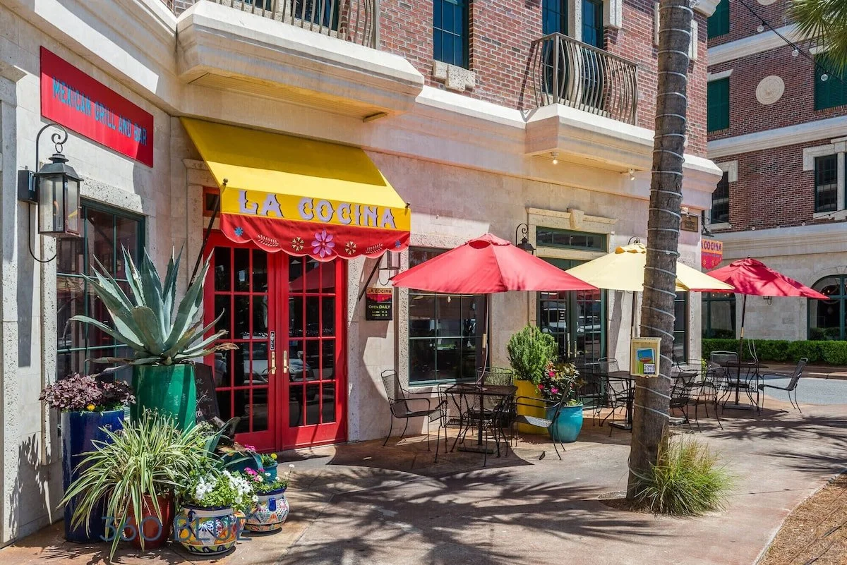 Exterior of a restaurant called LaCo Latin Costal Cuisine with red double doors, colorful awning, and outdoor seating with red and yellow umbrellas, potted plants, and a tree.