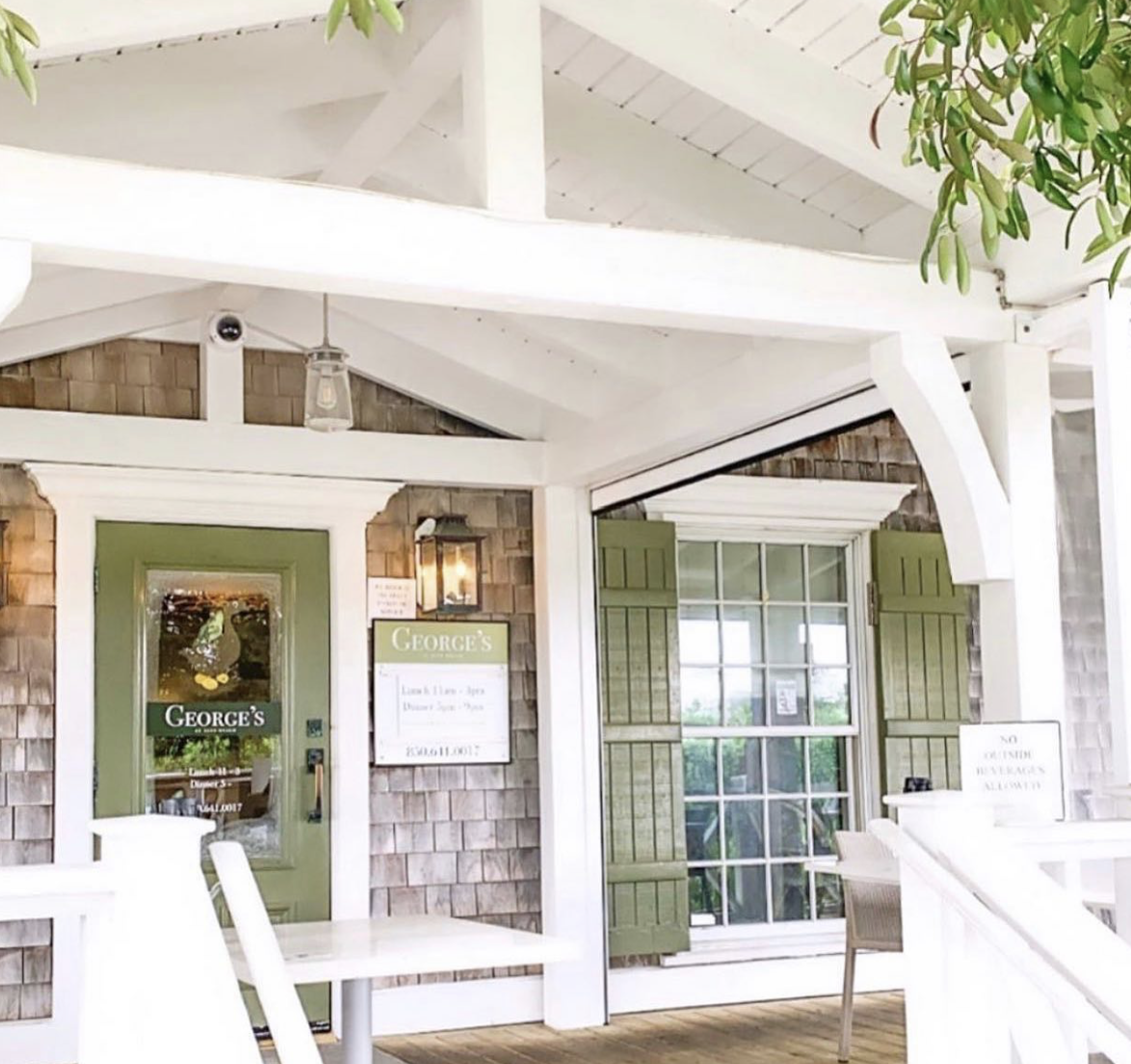 Exterior patio area of a restaurant called George's with a green door, window with green shutters, and outdoor tables and chairs, surrounded by a wooden exterior and greenery.
