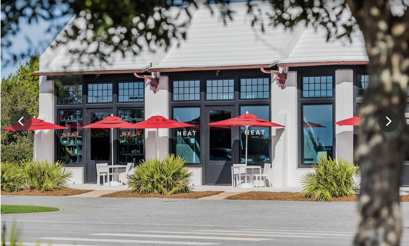 A bottle shop called NEAT. Modern cafe building with large glass windows, red umbrellas outside, white chairs, and green shrubs in the foreground.