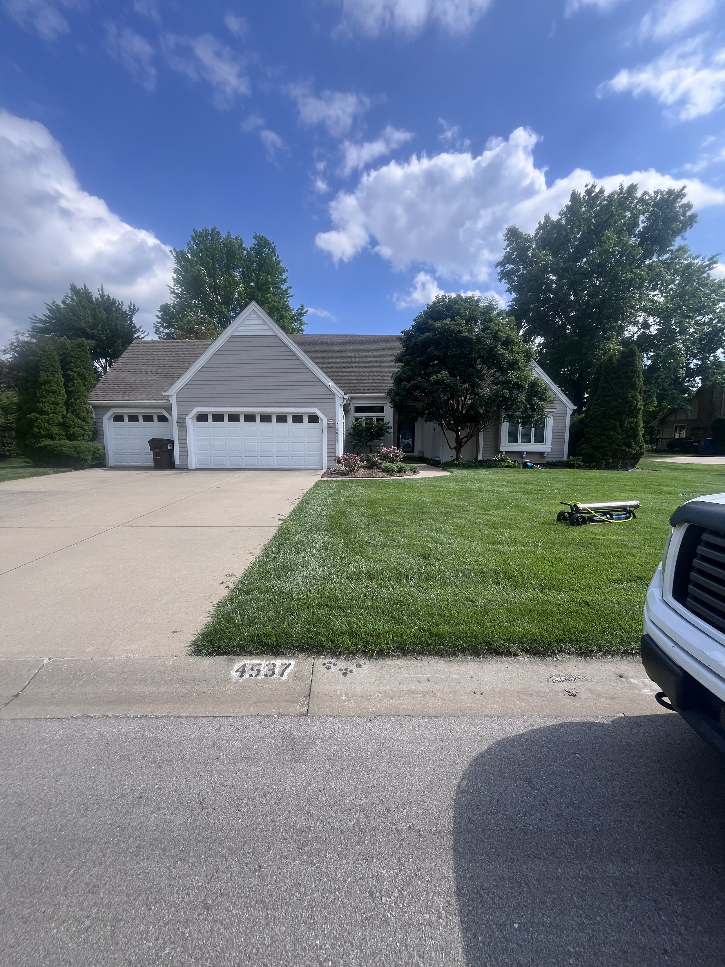 Front view of a white suburban house with a two-car garage, surrounded by a green lawn, trees, and shrubs on a sunny day with a blue sky and some clouds.