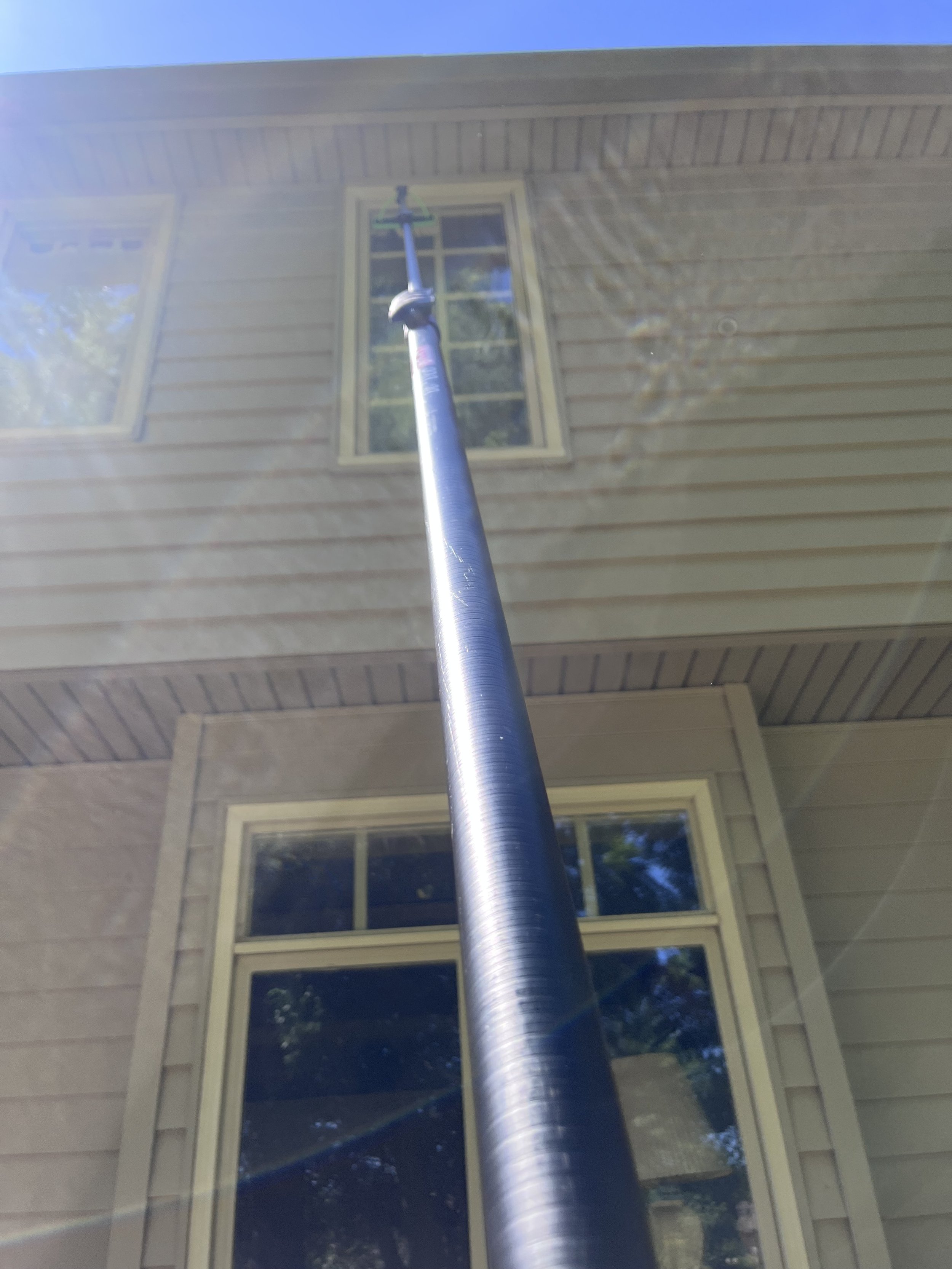 A tree trimming pole reaching up to a second story window of a beige house with vinyl siding, with sunlight creating lens flare.