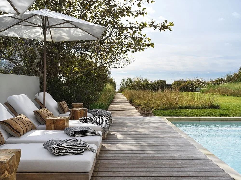 Poolside outdoor lounge area with white cushioned seats, striped towels, wooden side tables, large white umbrella, on a wooden deck next to a swimming pool, with a grassy landscape and trees in the background.