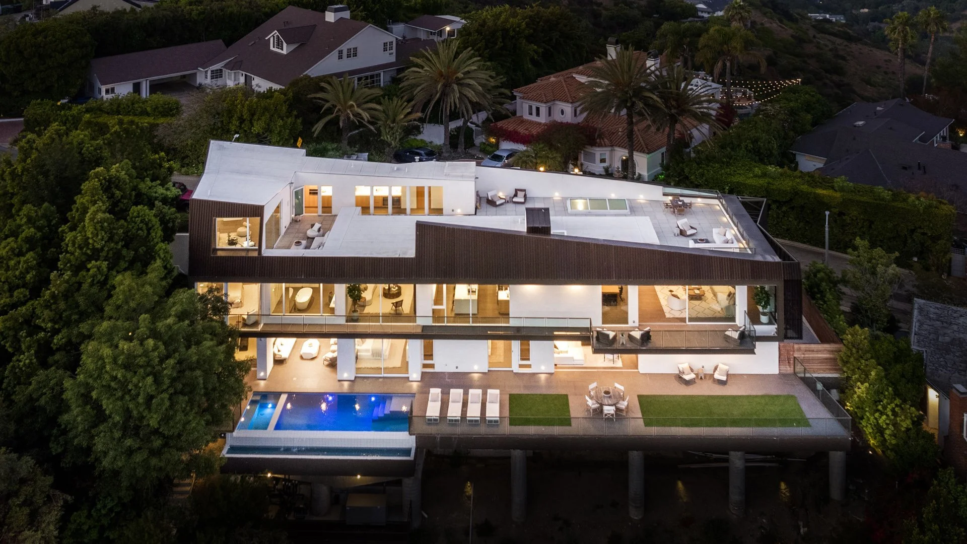A modern multi-story house with a rooftop deck, outdoor seating, a pool, and adjacent lawns, surrounded by trees and neighboring houses at dusk.
