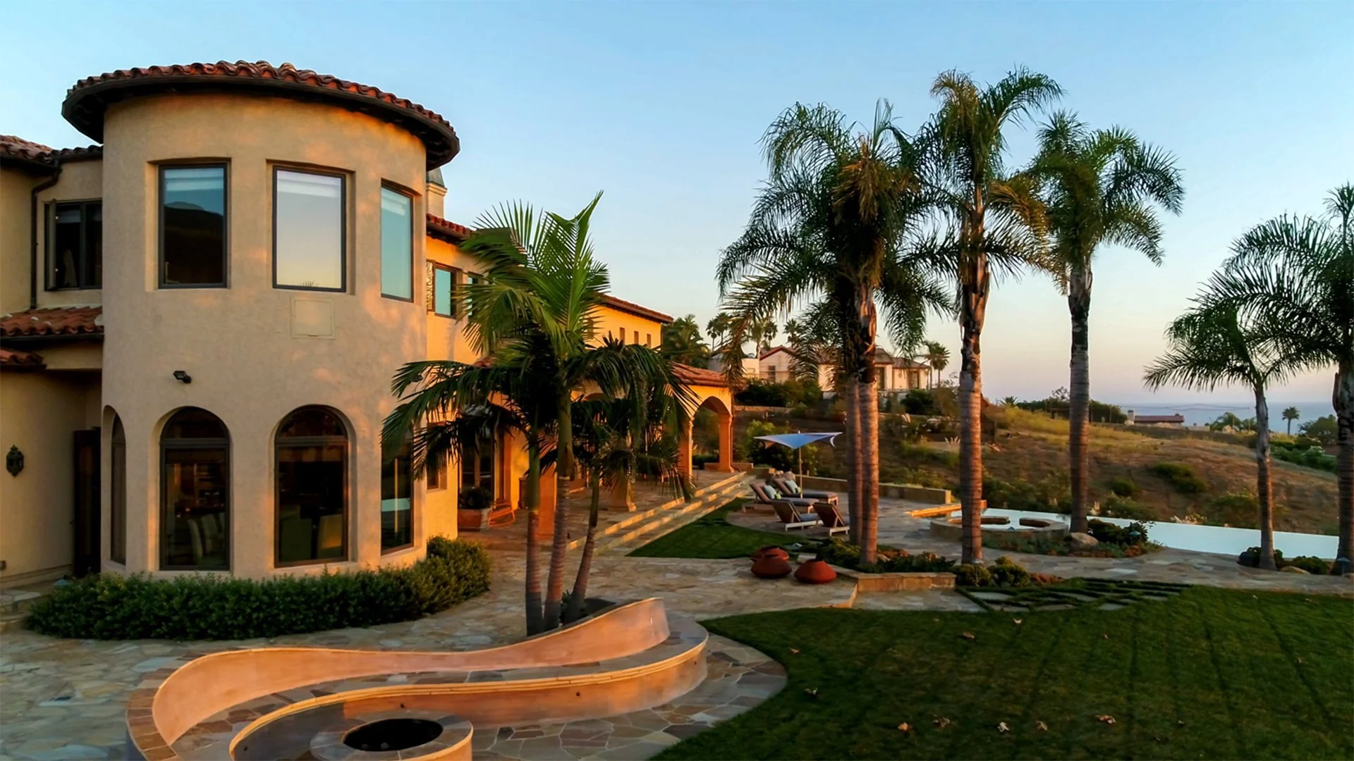 Luxury house with a curved tower and large windows, surrounded by palm trees, a patio with lounge chairs and an umbrella, and a hillside view at sunset.