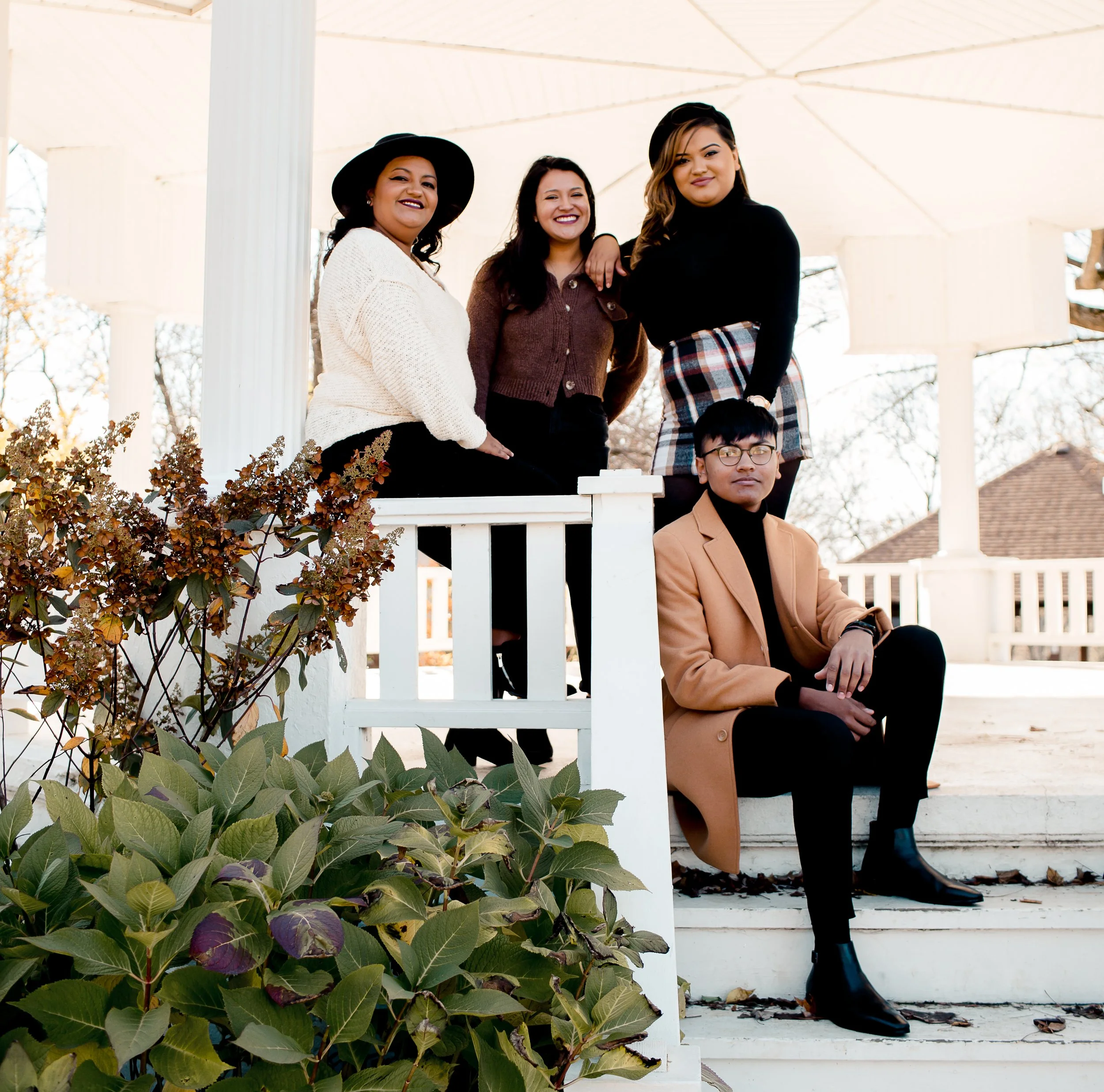 Group of four young adults, three women and one man, posing on the steps of a white gazebo outdoors with trees and a house in the background.