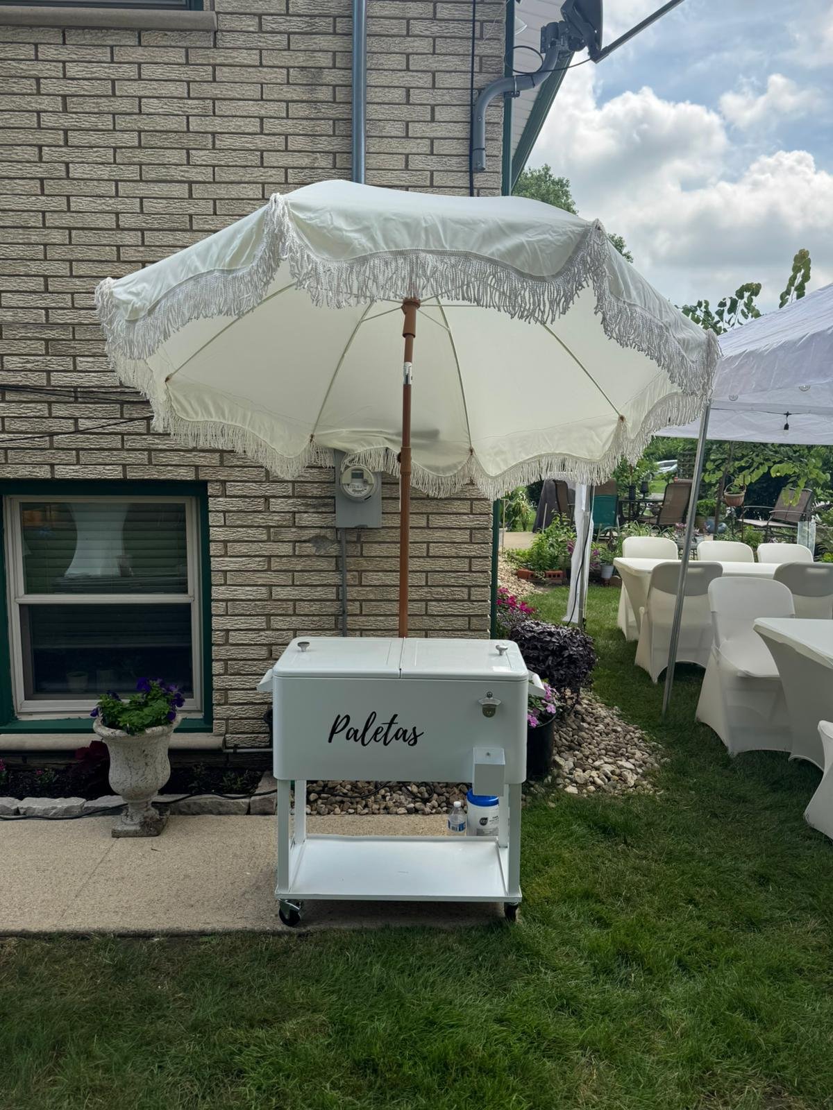 A white cart with the word 'Paletas' written on it, set outside under a large white umbrella with fringes. The cart is on wheels and located near a brick building with a window, on a grassy area with tables covered by white umbrellas in the backgroun
