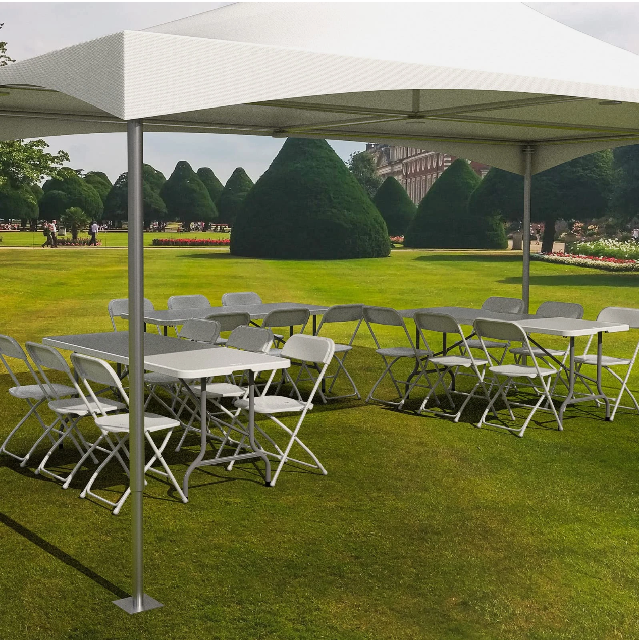 Empty white folding tables and chairs under a large white canopy on a well-manicured lawn with leafy green trees and trimmed bushes in the background.