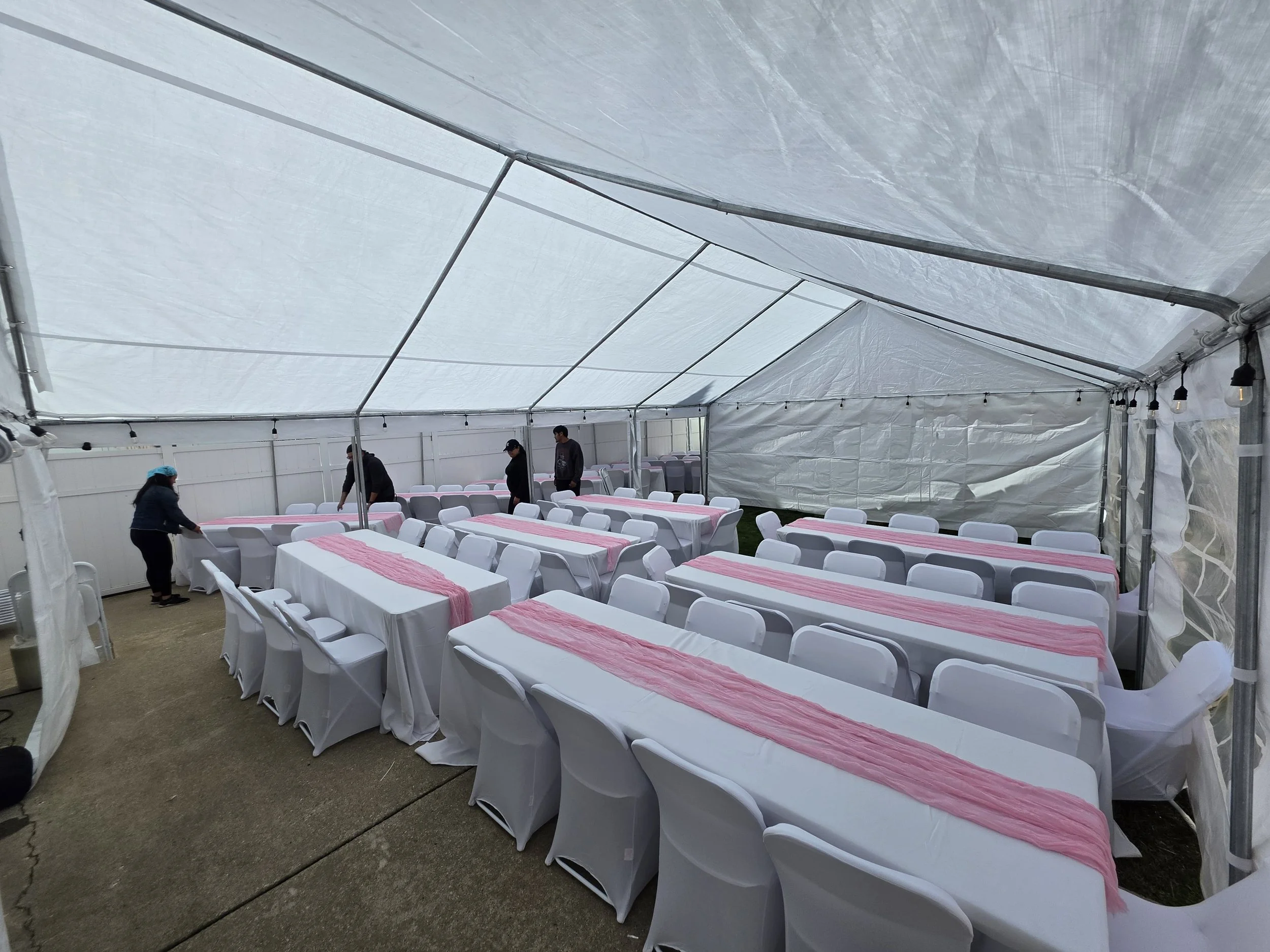 A large white event tent with tables covered in white tablecloths and pink runners. Several chairs are arranged around the tables, and a few people are inside preparing or setting up. String lights are hanging along the sides of the tent.