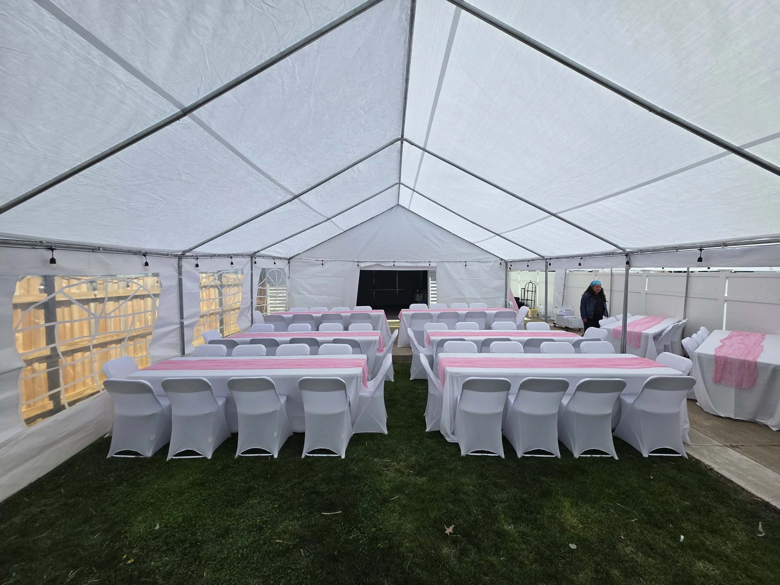 Inside a large white event tent with white chairs and tables covered in pink and white tablecloths, set up for a gathering or celebration, with string lights hanging from the ceiling.
