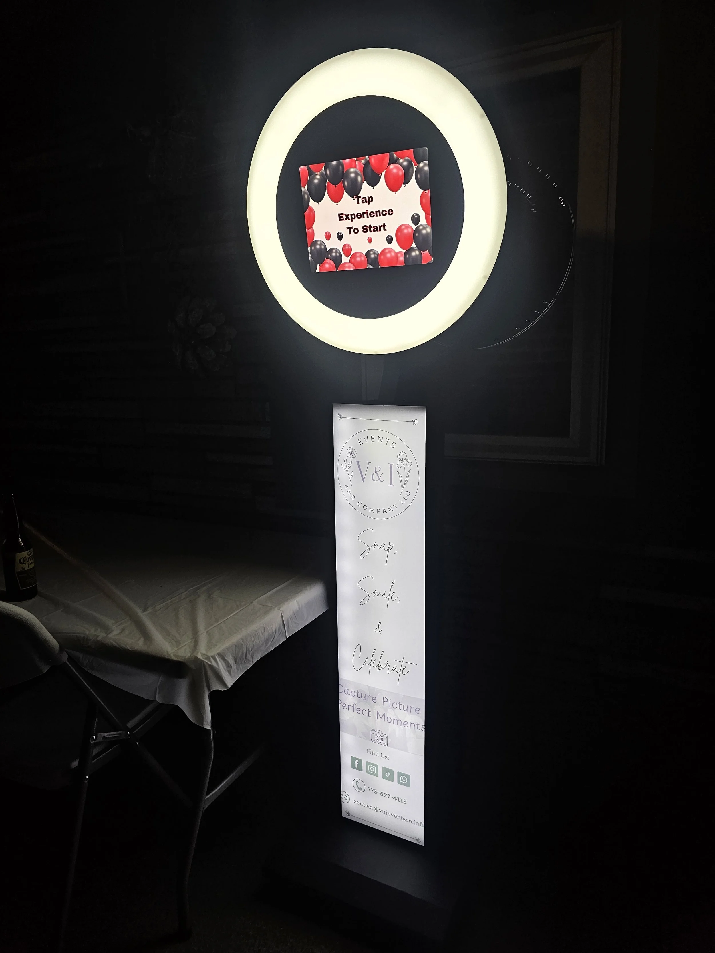 A promotional sign for an event with a black and red balloon background and white text that reads, "Tap Experience To Start." Next to the sign, a vertical candle is sitting on a table covered with a white tablecloth, with a drink bottle positioned to