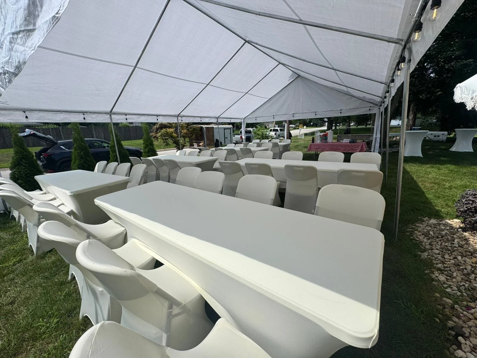 Long tables with white chairs arranged under a large white event tent outdoors on a grassy area, with some cars and trees visible in the background.