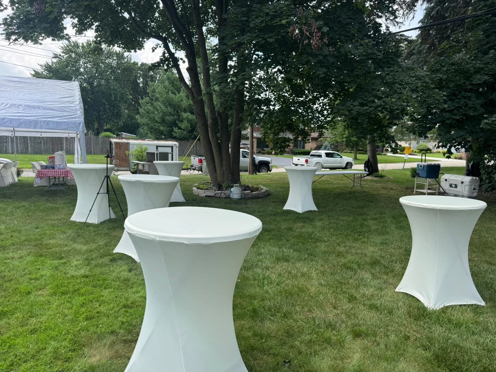Outdoor event setup with white high-top tables on a grassy lawn, a white tent on the left, a tree, and some vehicles in the background.