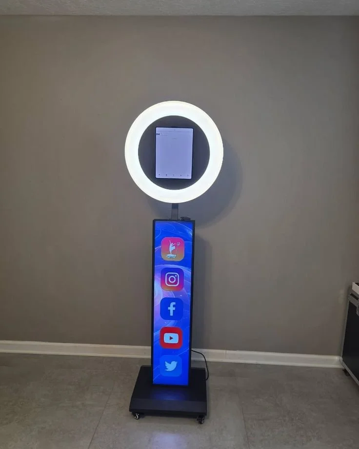 A digital social media kiosk featuring a vertical display with icons for TikTok, Instagram, Facebook, YouTube, and Twitter, placed on a mobile stand against a plain wall with a ring light and a tablet above.