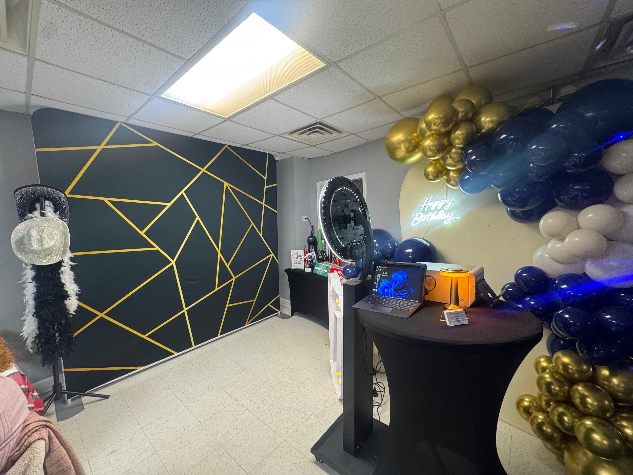 Party decorations with gold, black, blue, and white balloons arranged in a cluster, a black and yellow geometric patterned wall, and a neon sign that says 'Happy Birthday' in white.
