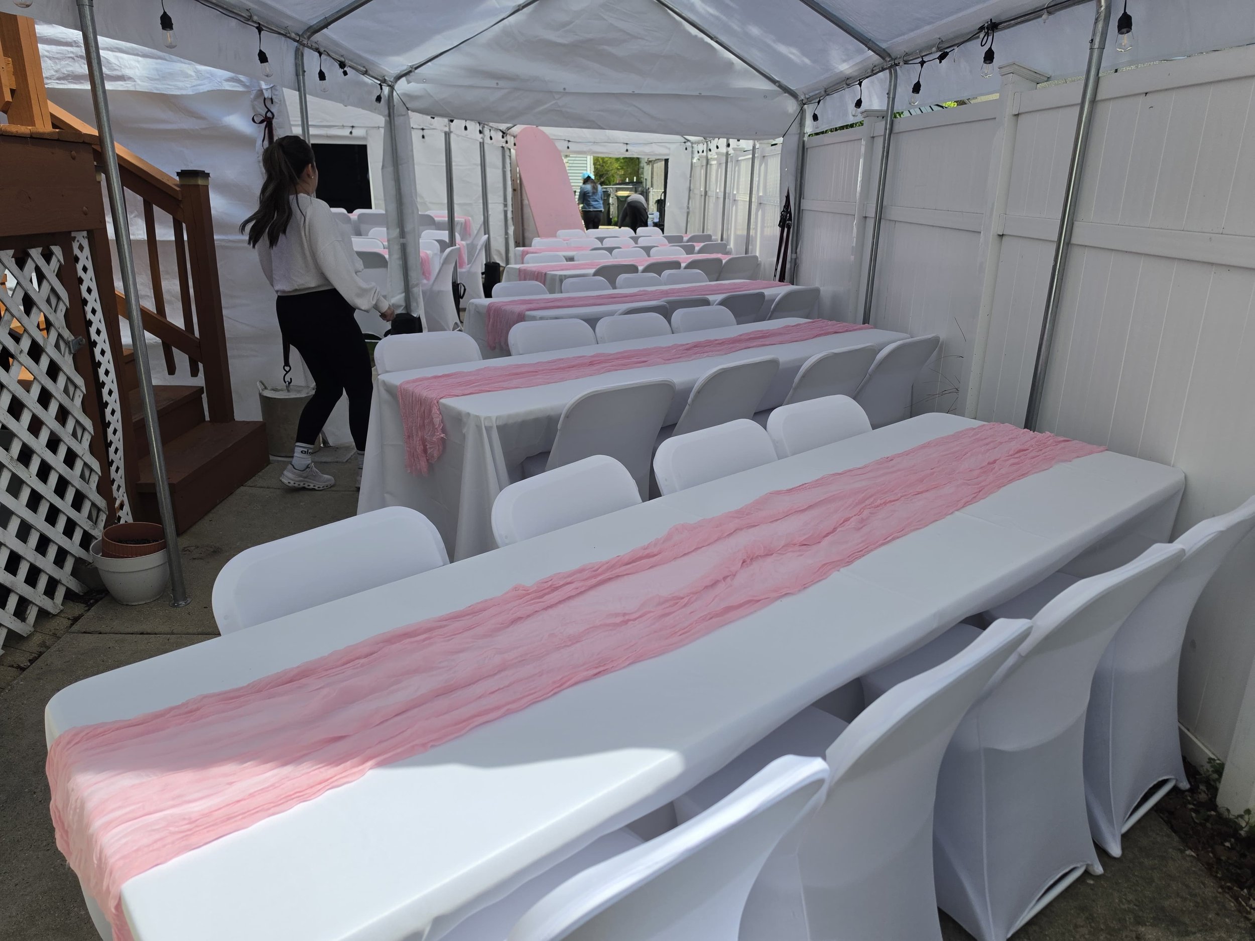Empty outdoor event tent with rows of white chairs covered with pink fabric table runners. A woman is standing near the entrance, and there are string lights hanging from the ceiling.