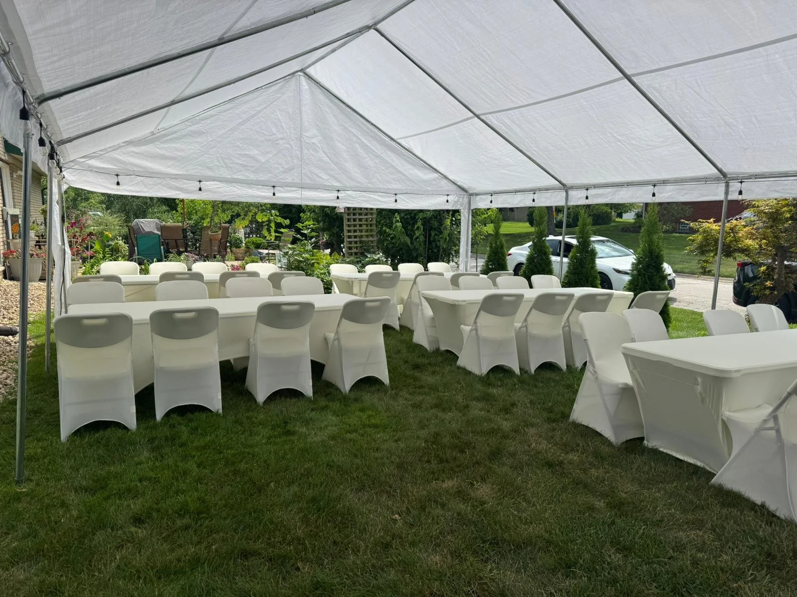 Outdoor event setup with white tables, chairs, and canopy tent in a garden area.