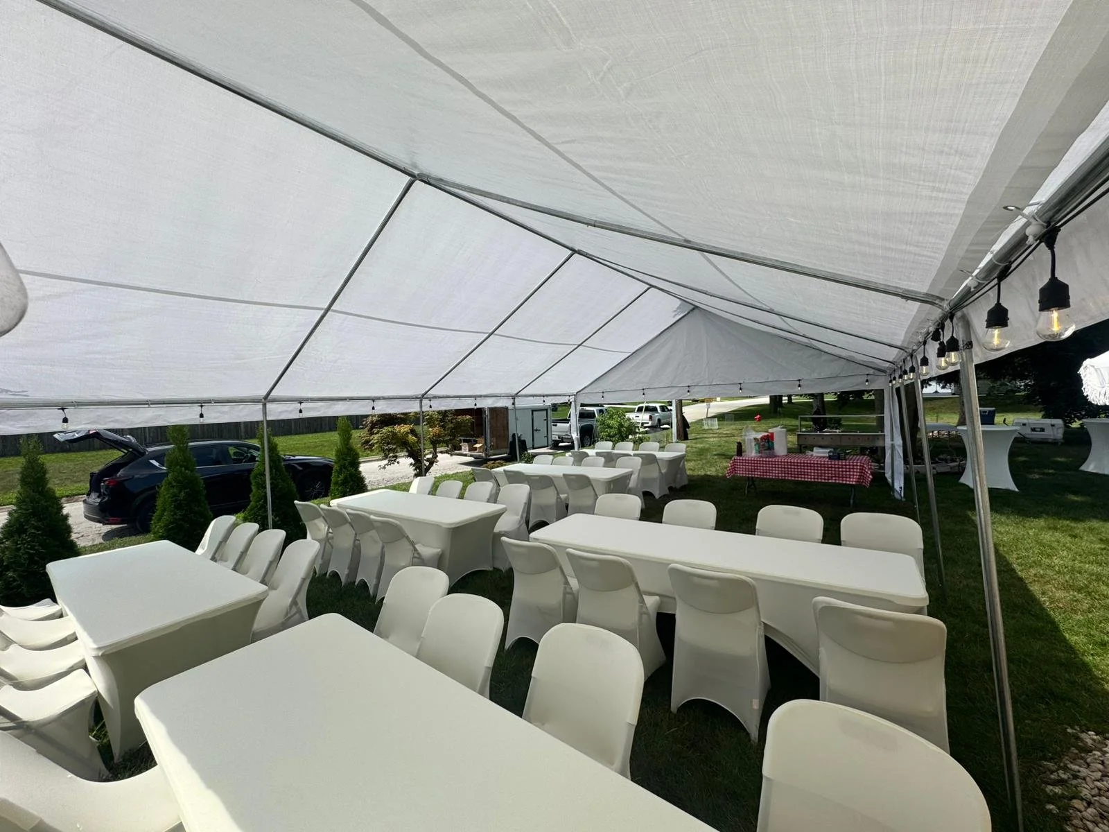 Empty outdoor event tent with white tables and chairs, decorated with string lights, set on grass during the day.