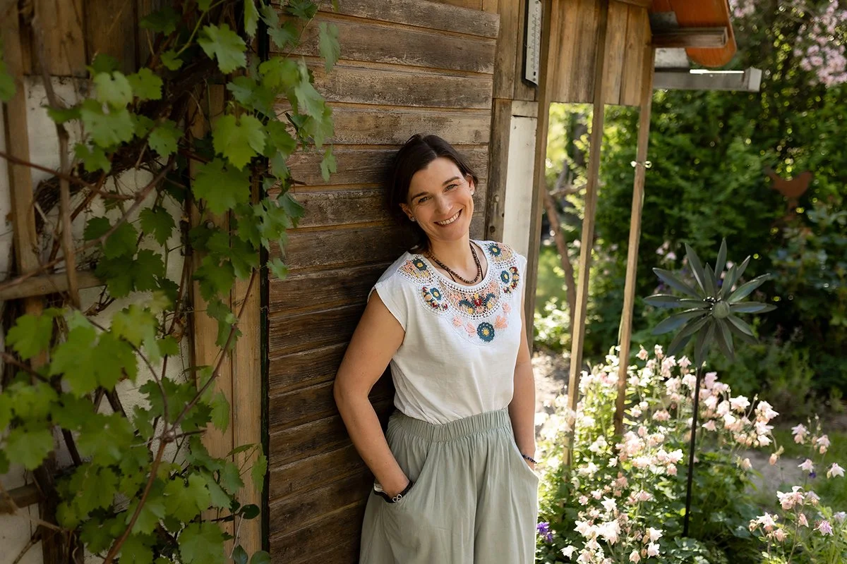 Junge Frau mit dunklen Haaren, lächelnd, steht an einer Holzvertäfelung vor einem Garten mit Blumen und grünen Pflanzen, trägt ein weißes Oberteil mit buntem Stickmuster und eine hellgraue Hose.