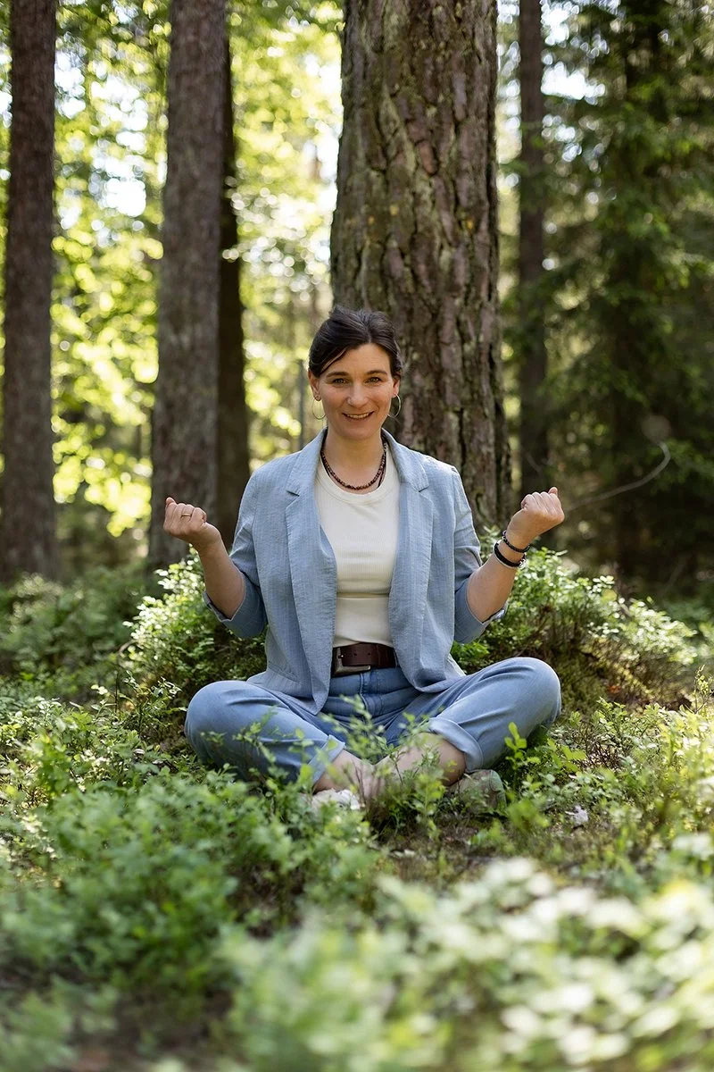 Eine Frau in Business Kleidung sitzt im Wald auf dem Boden, umgeben von Grün und Bäumen, in entspannter Pose. Sie gibt einen Kurs zum Thema Entspannung. Progressive Muskelentspannung, Progressive Muskelrelaxation, PME