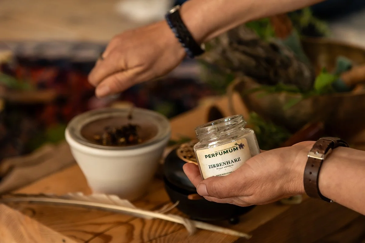 Anja Haselhofer zeigt eine Räuchermischung und arbeitet mit einem Stövchen auf einem Holztisch, umgeben von Pflanzen und Naturmaterialien.