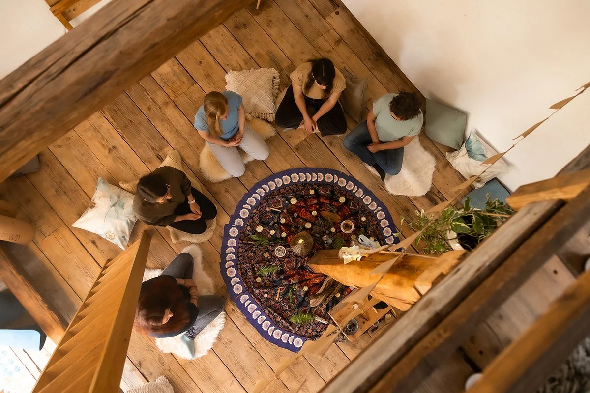 Fünf Frauen sitzen in gemütlicher Runde auf Bodenkissen um einen liebevoll gestalteten Teppich mit Kerzen und Naturdekoration. Die harmonische Atmosphäre schafft Raum für Entspannung, Austausch und gemeinsame Zeit unter Freundinnen.
