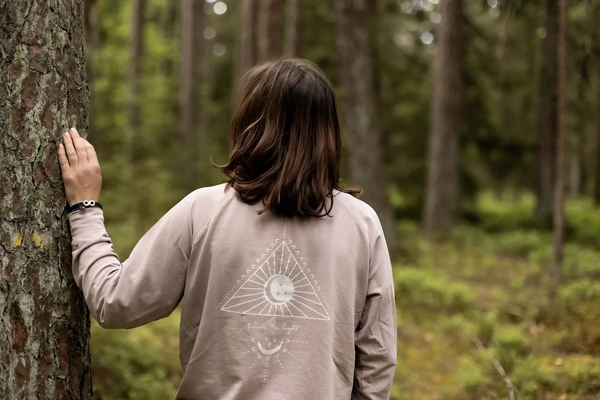 Frau in einem Wald mit ihrer Rückseite zum Betrachter, trägt ein hellgraues Sweatshirt mit einem mystischen Design auf dem Rücken.