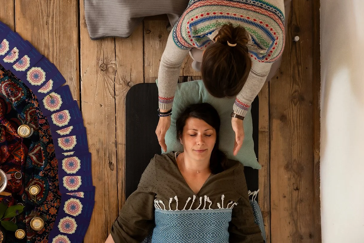 Eine Frau liegt auf einer Yogamatte mit geschlossenen Augen, während eine andere Frau sie mit Energiearbeir und Aromaölen behandelt. Der Raum hat Holzboden und schön eingerichtet. 1:1 Begleitung, Aromaöle, Reki