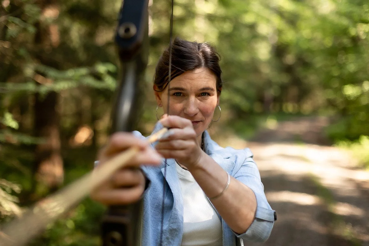 Eine Frau in einem Wald beim Bogenschießen, die den Bogen spannt und zielt. Fokus und Klarheit.