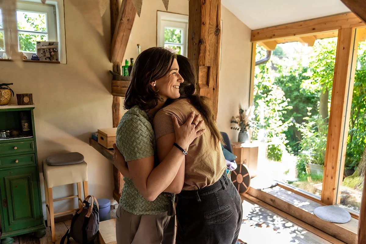Zwei Frauen umarmen sich in einem hellen Raum mit Holzbalken und genießen einen wertvollen Moment der Nähe und Begegnung.