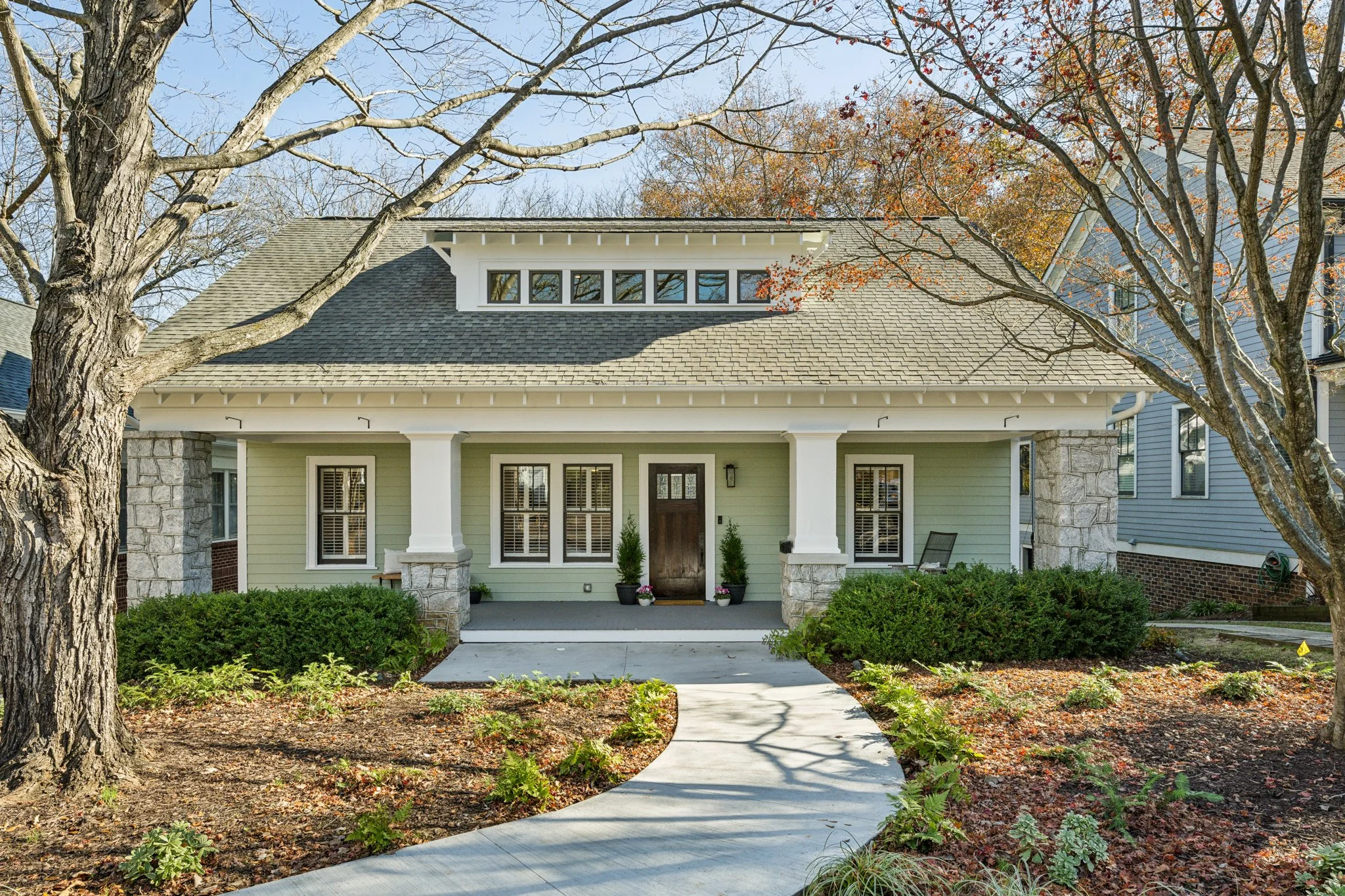 Front view of a house with light green siding, stone accents, and a front porch with columns, surrounded by trees and landscaped yard with a curved walkway.