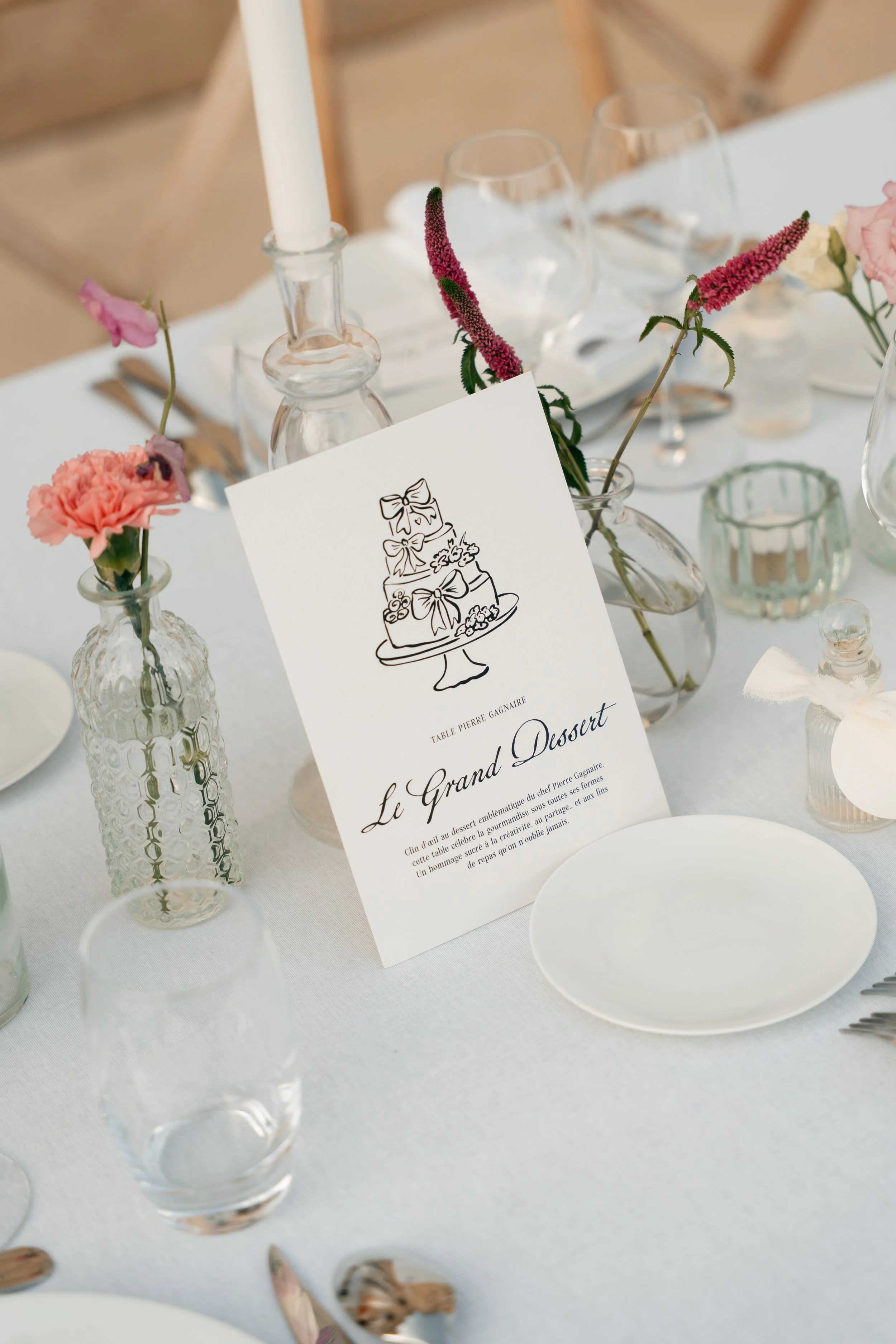 Papeterie de mariage J&A : affiche de table avec illustration d'un gâteau décoré, placée sur une table élégante avec des verres, vases en verre avec des fleurs, et d'autres éléments de décoration pour un événement.