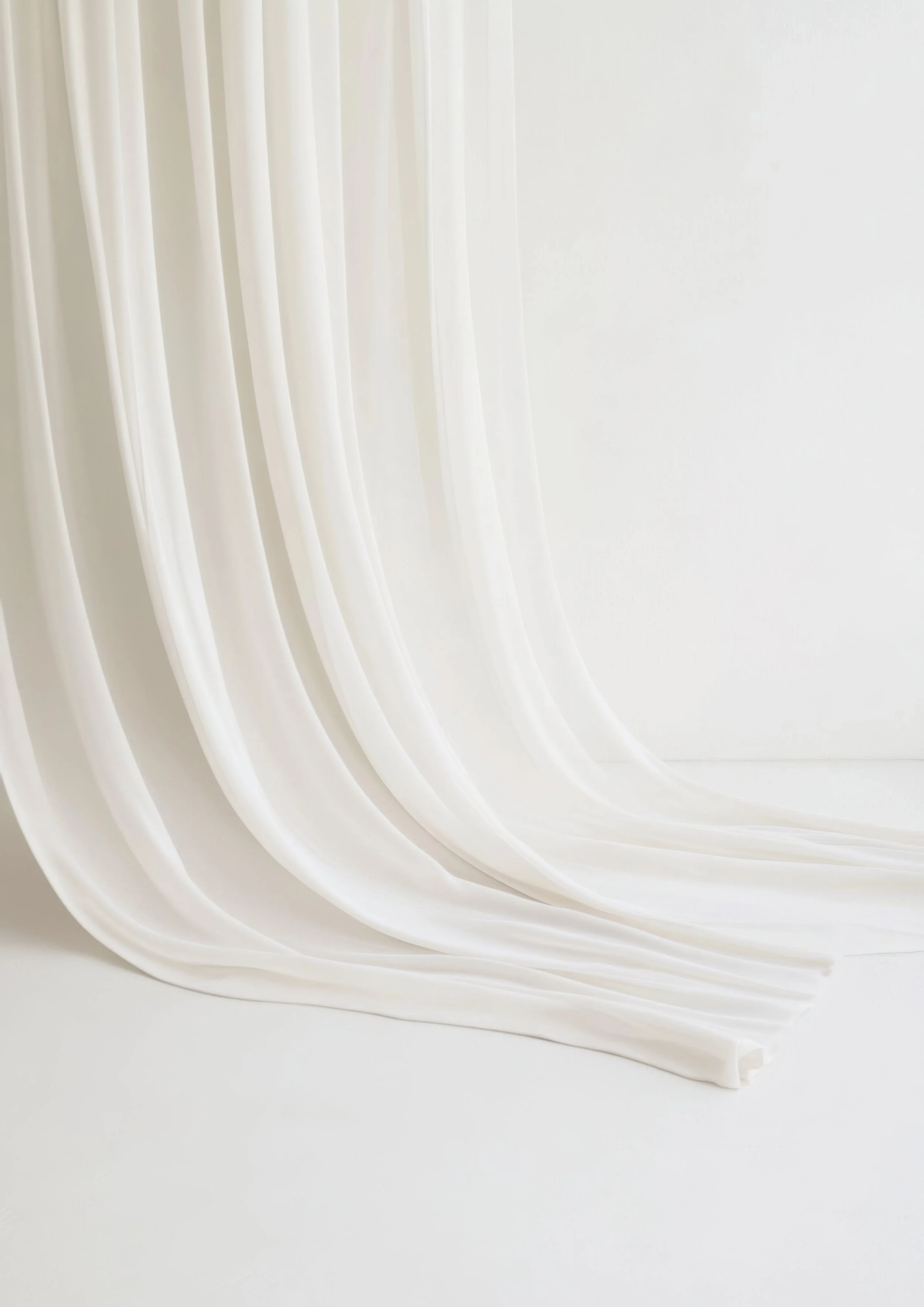 Des rideaux en tissu blanc suspendus avec des plis, tombant sur un sol blanc.