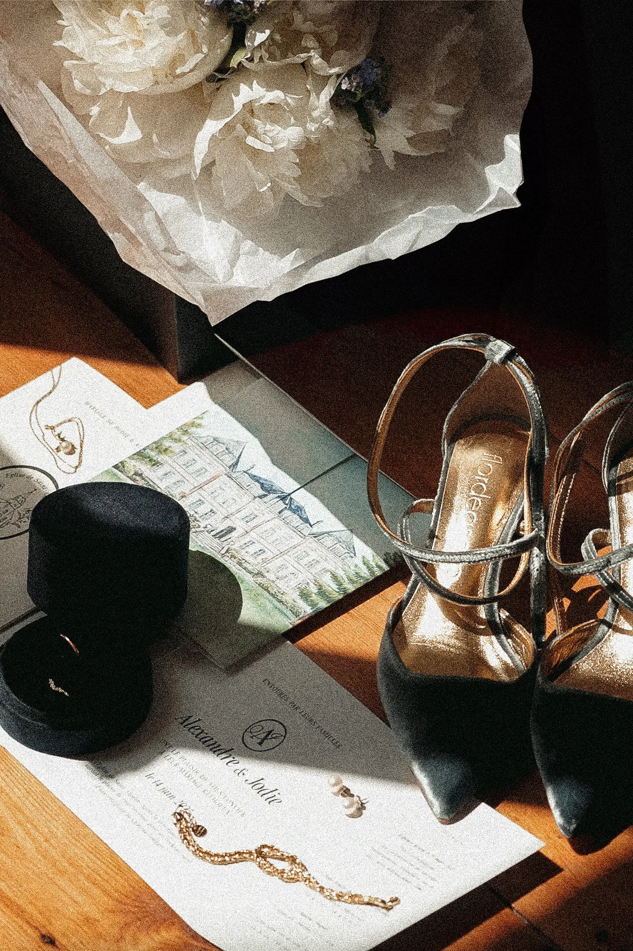 Papeterie de mariage, une paire de chaussures à talons, une boîte à bijoux, des bijoux, et des fleurs. Luminosité chaleureuse.