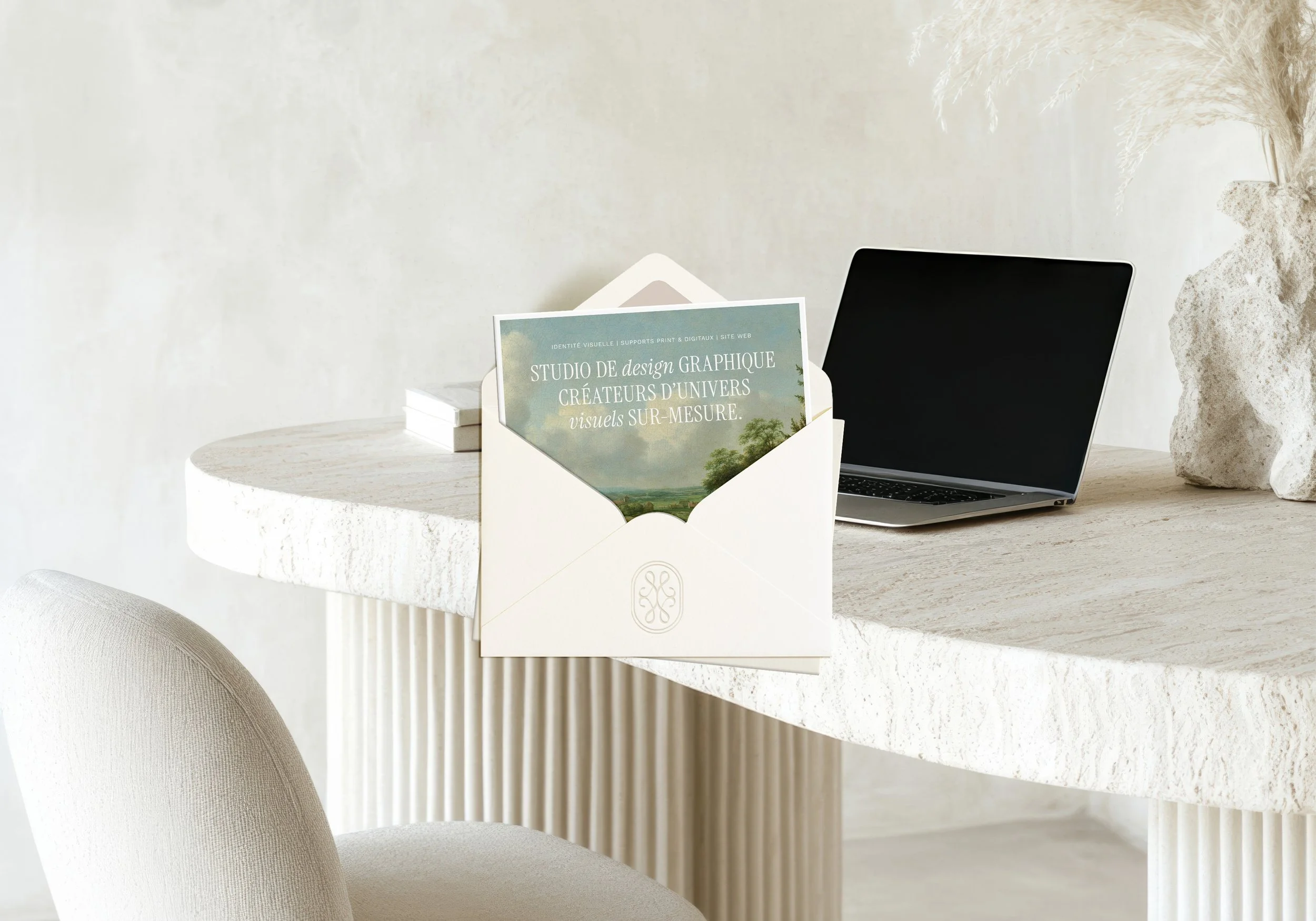 Carte dans une enveloppe avec inscrit : "Studio de design graphique créateur d'univers visuels sur-mesure ». Fond de l’image représentant un bureau avec un ordinateur portable, des livres et une chaise dans un intérieur minimaliste.