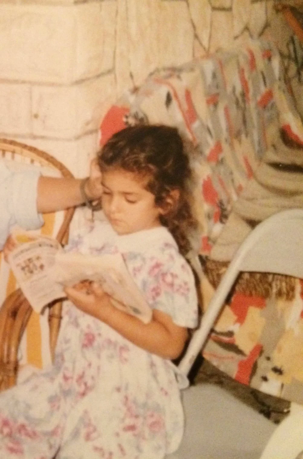 A young girl with curly hair sitting on a bed, reading a book.