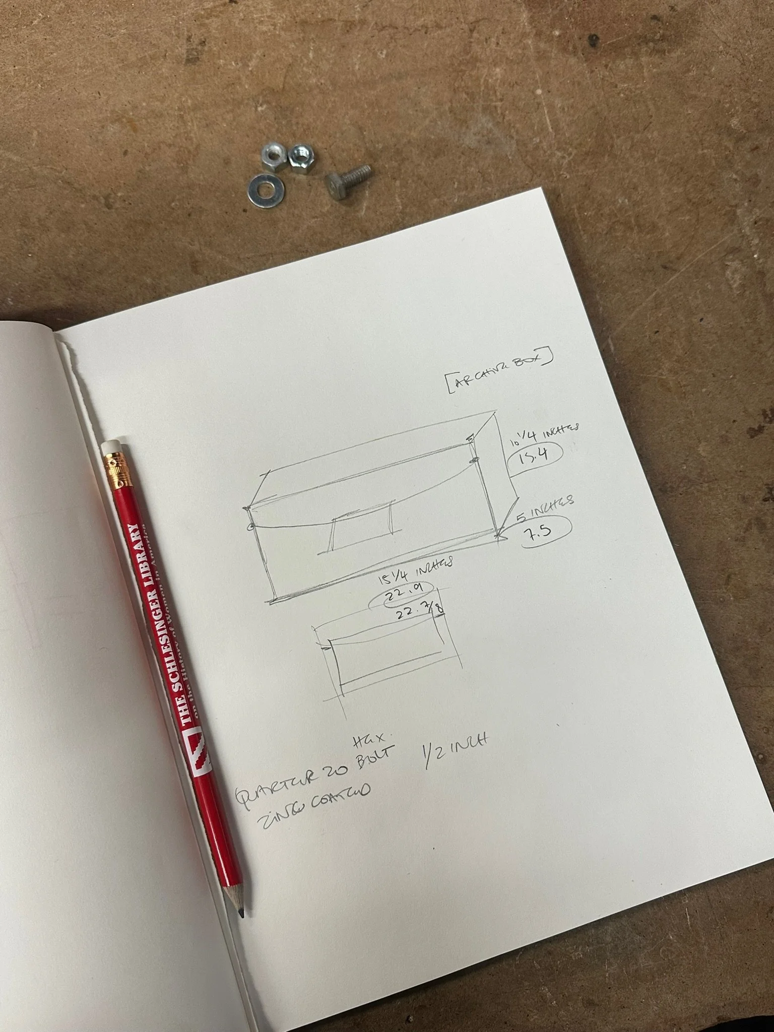 Sketch of a rectangular box with measurements, a notepad, a red pen, and some screws on a wooden surface.