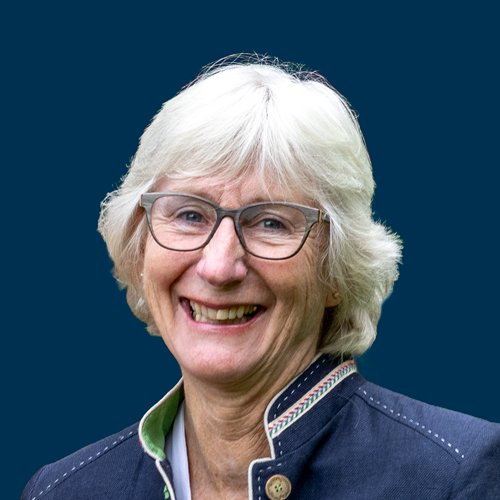 A smiling woman with short white hair and glasses, wearing a dark navy blazer with a high collar and a light green shirt underneath, against a solid dark blue background.