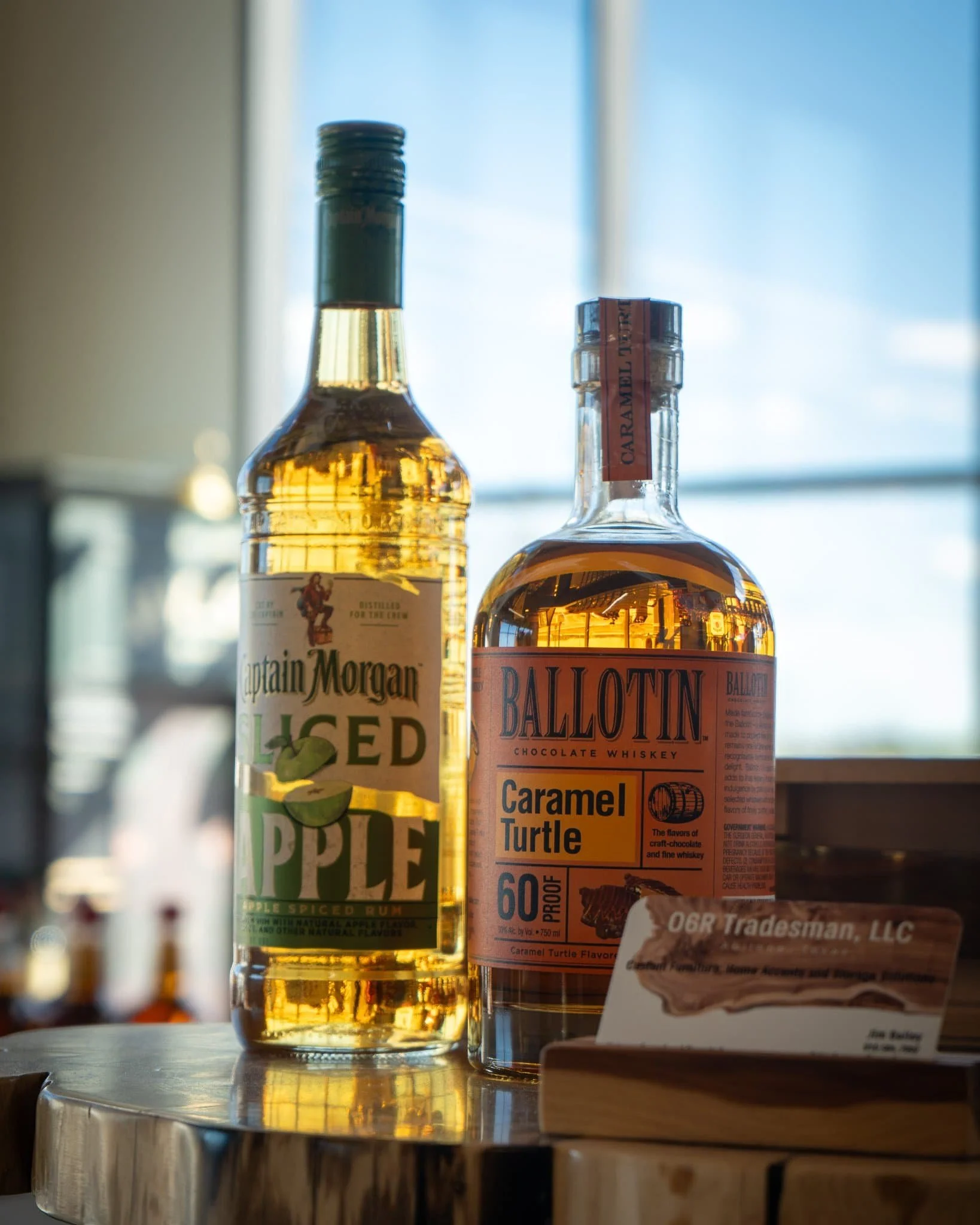 Two bottles of alcohol, one labeled Captain Morgan Spiced Apple Rum and the other Ballotin Chocolate Whiskey Caramel Turtle, on a reflective wooden surface with a blurred background of a window and other bottles.
