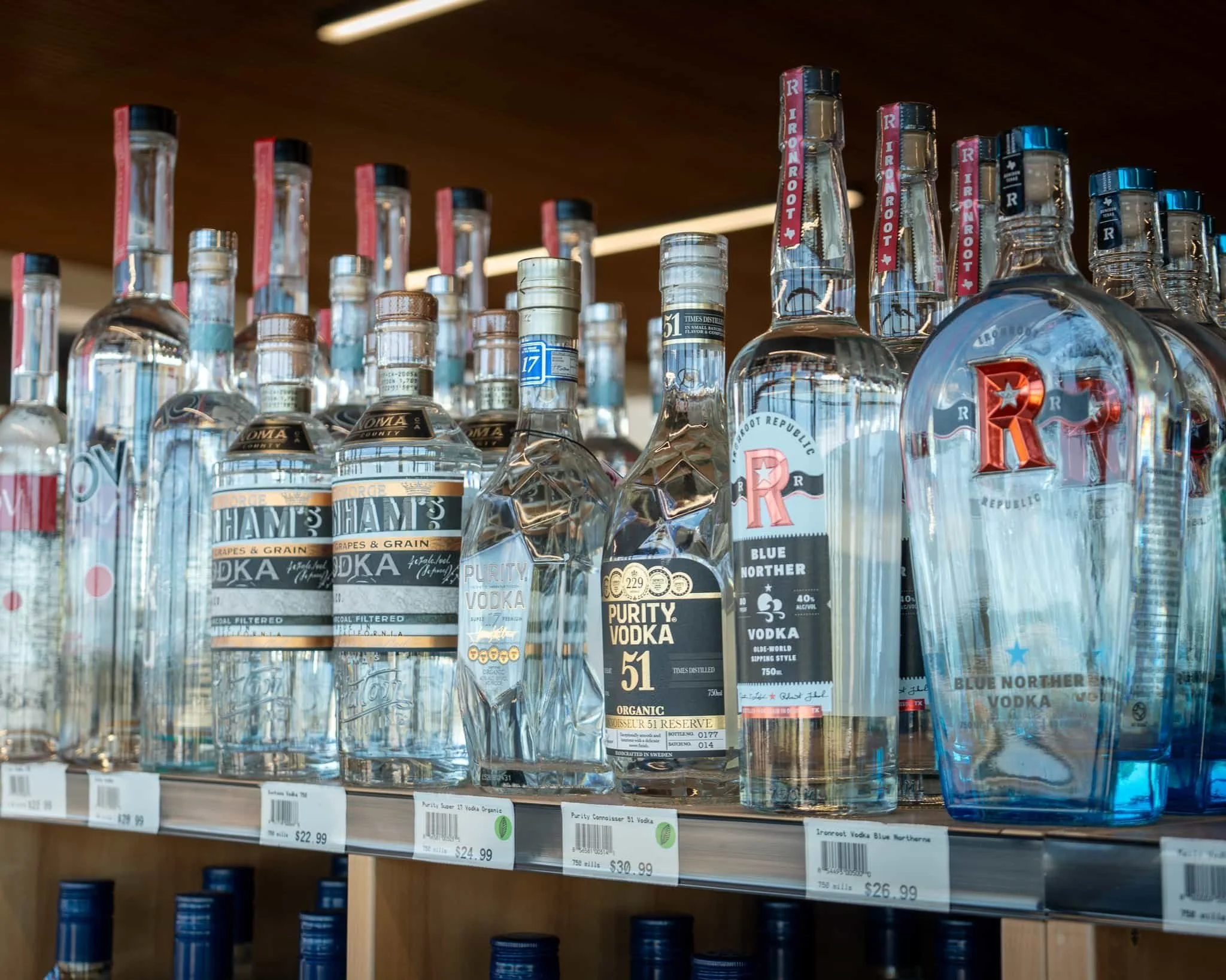 Shelf of various brands of vodka bottles, including Purity, Ham's, and Ironroot, with price tags underneath.