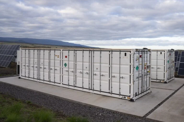 Two white container units with security latches on a concrete pad, surrounded by solar panels in a rural landscape under cloudy skies.