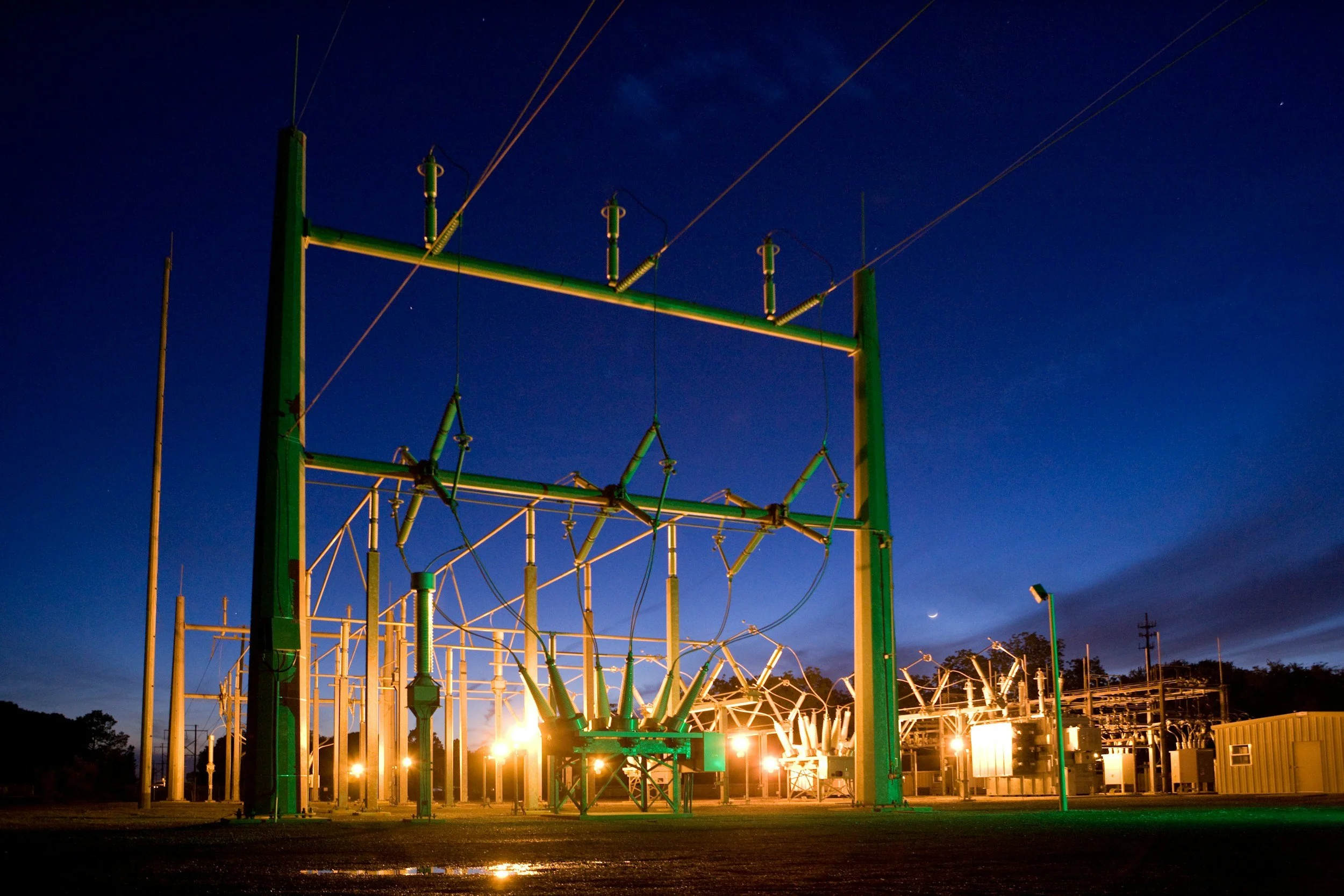 An electric power substation at night with illuminated transformers, insulators, and power lines against a dark blue sky with a crescent moon.