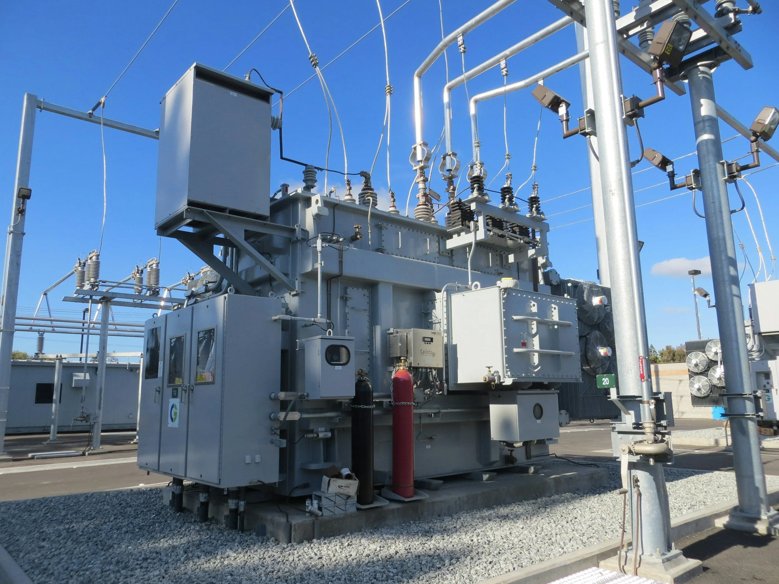 Electric substation equipment including transformers, insulators, and power lines against a clear blue sky.