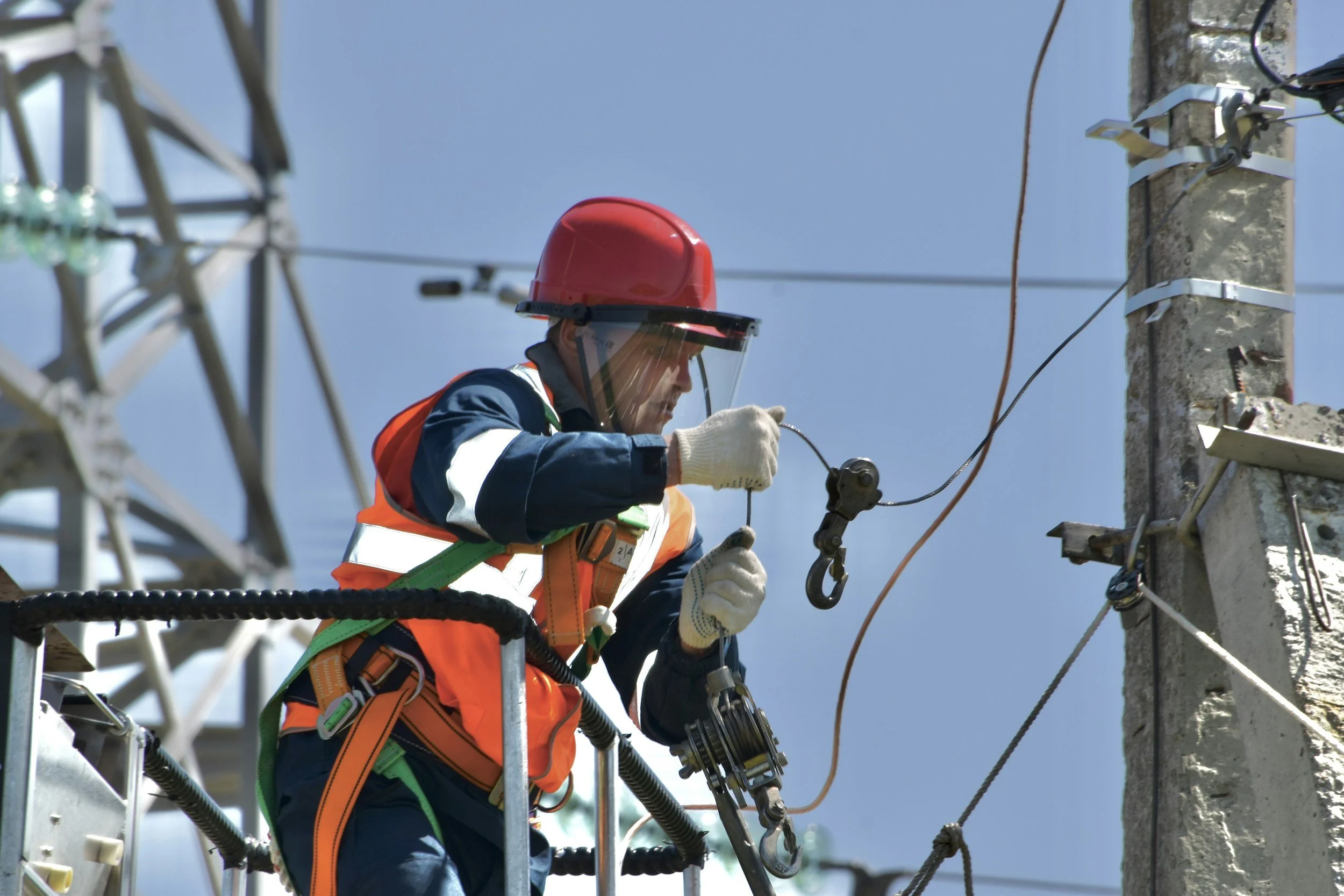 A worker in safety gear, including a red helmet, face shield, orange safety vest, and gloves, is inspecting or working on electrical wires attached to a utility pole from a raised platform against a blue sky background.